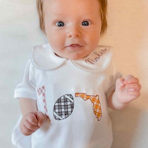 May include: A white baby onesie with a Peter Pan collar. The onesie features embroidered designs of a football, the state of Florida, and the word "Noles". The football and Florida designs have plaid patterns.