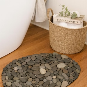 May include: Oval-shaped stone bath mat made of various sized gray and tan river rocks. A woven basket with a book titled "TUSCANY MARVEL" and a white towel are in the background. A white bathtub and a beige towel are also visible.