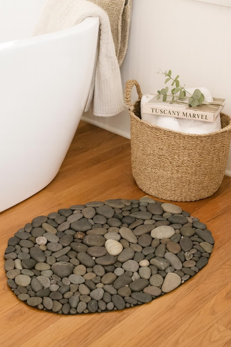 May include: Oval-shaped stone bath mat made of various sized gray and tan river rocks. A woven basket with a book titled "TUSCANY MARVEL" and a white towel are in the background. A white bathtub and a beige towel are also visible.