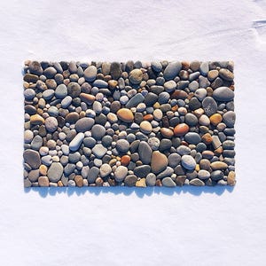 Pu&ograve; includere: Un tappetino rettangolare composto da numerose pietre di fiume lisce e arrotondate in varie tonalit&agrave; di grigio, beige, marrone e bianco. Le pietre sono disposte strettamente in un motivo piatto e denso, creando una superficie strutturata.