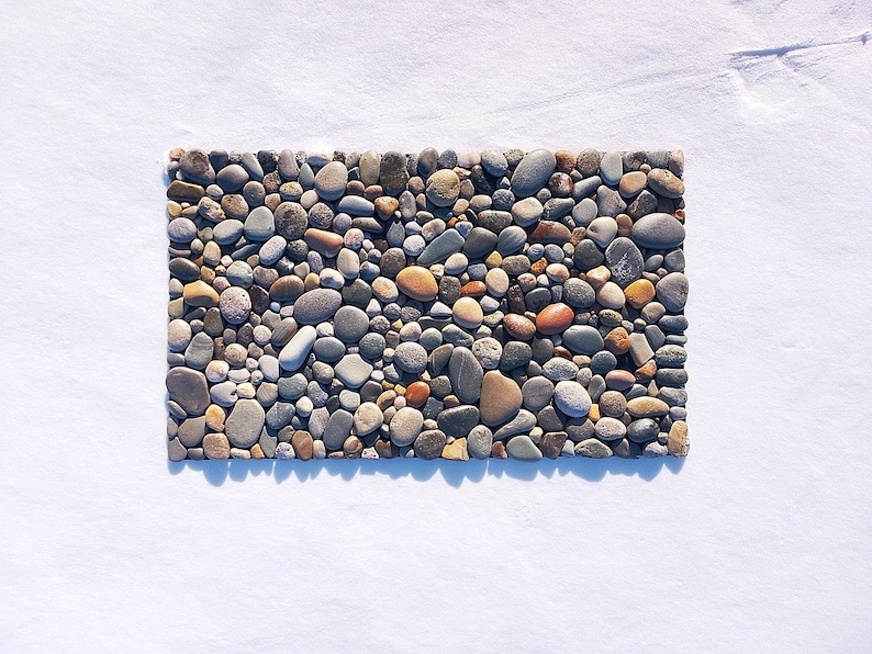 Pu&ograve; includere: Un tappetino rettangolare composto da numerose pietre di fiume lisce e arrotondate in varie tonalit&agrave; di grigio, beige, marrone e bianco. Le pietre sono disposte strettamente in un motivo piatto e denso, creando una superficie strutturata.