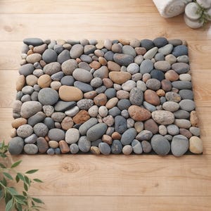 Puede incluir: Una alfombra de baño rectangular hecha de piedras de río lisas y redondeadas en tonos grises, marrones y beige. Las piedras están dispuestas juntas sobre un respaldo negro, creando una superficie natural y texturizada. La alfombra descansa sobre un suelo de madera clara.