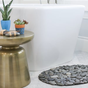 May include: A bathroom scene featuring a gold side table with potted plants and decorative stones. A white bathtub is in the background, and a gray stone pebble bath mat is on the floor. The scene evokes a spa-like atmosphere.