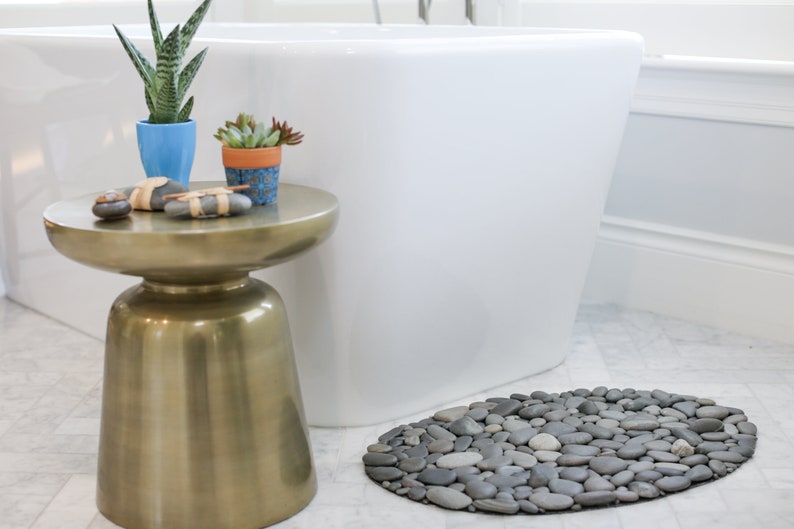 May include: A bathroom scene featuring a gold side table with potted plants and decorative stones. A white bathtub is in the background, and a gray stone pebble bath mat is on the floor. The scene evokes a spa-like atmosphere.