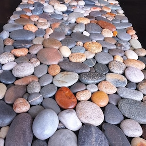 Pu&ograve; includere: Un mosaico di ciottoli lisci e arrotondati in vari colori, tra cui grigio, marrone, arancione e bianco. Le pietre sono disposte vicine, creando una superficie strutturata. L'effetto complessivo &egrave; naturale e organico.