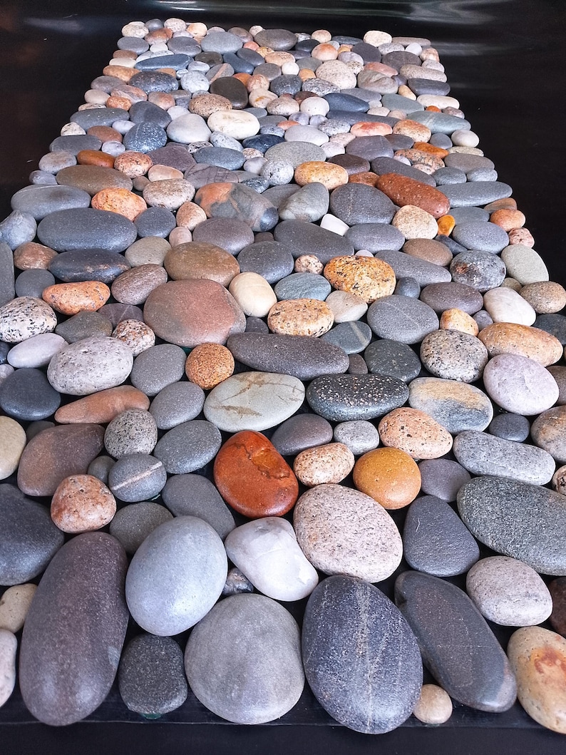 Pu&ograve; includere: Un mosaico di ciottoli lisci e arrotondati in vari colori, tra cui grigio, marrone, arancione e bianco. Le pietre sono disposte vicine, creando una superficie strutturata. L'effetto complessivo &egrave; naturale e organico.