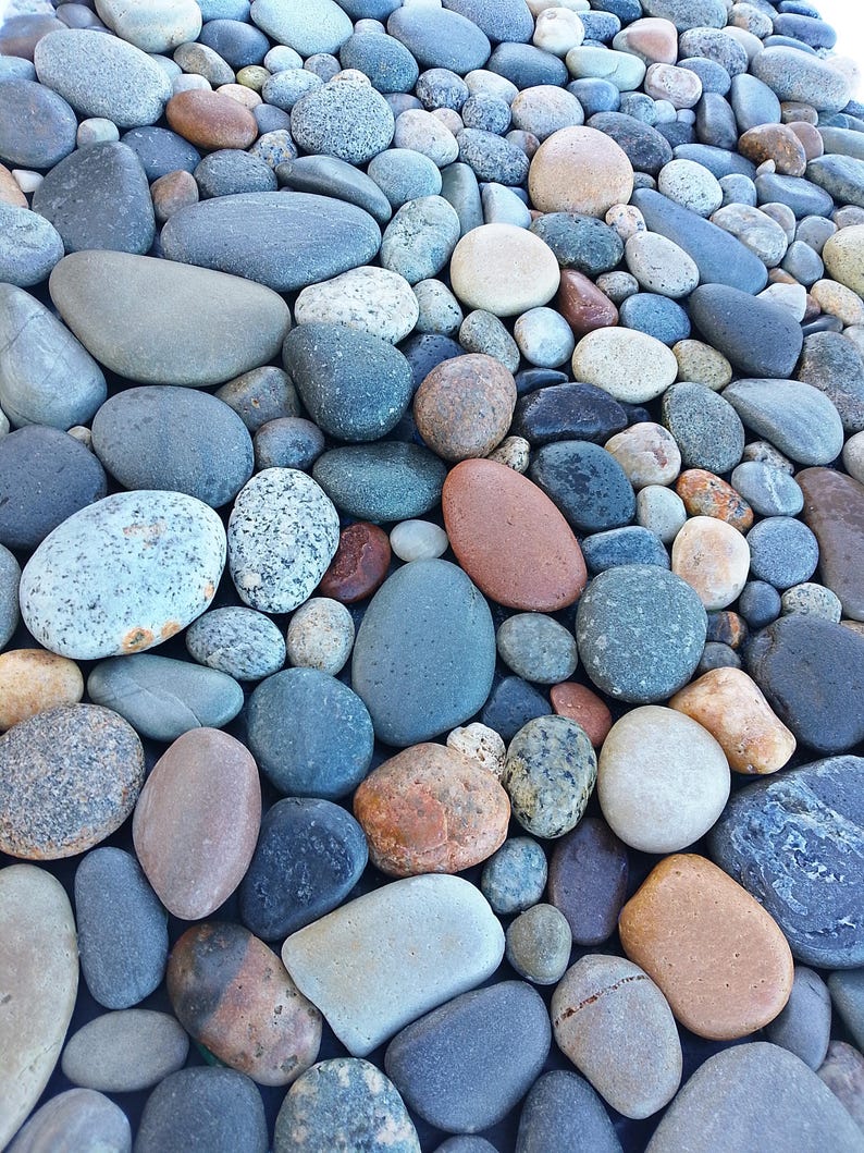 Pu&ograve; includere: Una collezione di pietre lisce e arrotondate in varie dimensioni e colori, tra cui sfumature di grigio, blu, marrone e beige. Le pietre sono strettamente raggruppate, creando una superficie naturale e strutturata.