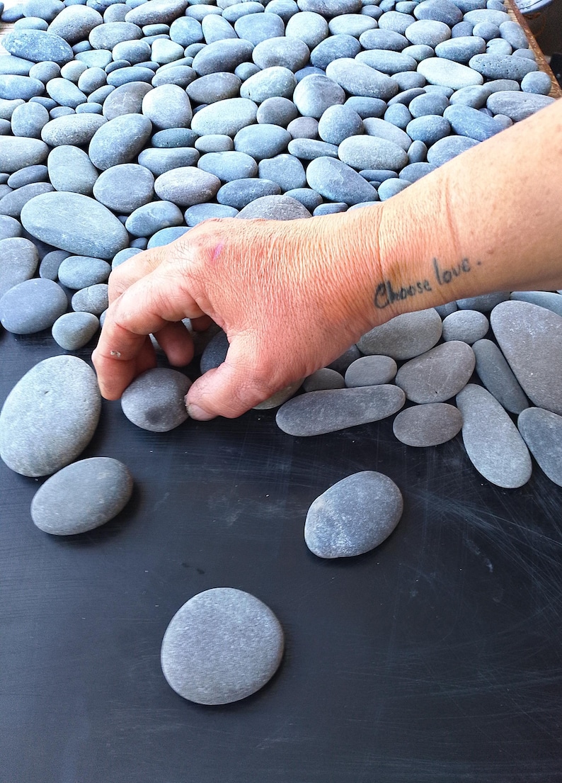Pu&ograve; includere: Disposizione di ciottoli di fiume grigi e lisci. Una mano posiziona una pietra su una superficie scura. Sul polso &egrave; tatuata la scritta "Choose love". Le pietre variano per dimensioni e forma, creando un'estetica naturale e organica.