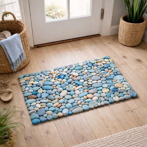 Pu&ograve; includere: Un tappetino rettangolare composto da pietre lisce e arrotondate nei toni del blu, grigio e beige. Il tappeto &egrave; posizionato su un pavimento in legno chiaro vicino a una porta bianca e un cesto di vimini.