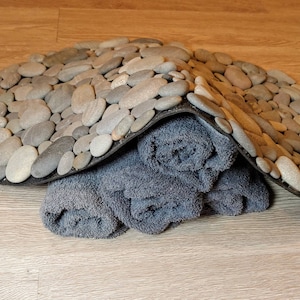 May include: A gray stone pebble bath mat with a black border, partially covering rolled-up gray towels. The pebbles are various sizes and shades of gray. The towels are a darker gray color.
