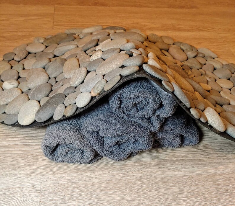May include: A gray stone pebble bath mat with a black border, partially covering rolled-up gray towels. The pebbles are various sizes and shades of gray. The towels are a darker gray color.