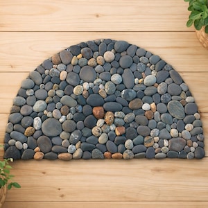 Può includere: Un tappetino in pietra a forma di semicerchio composto da ciottoli grigi, marroni e beige di varie dimensioni. Il tappetino è su una superficie di legno marrone chiaro, con piante in vaso sullo sfondo.