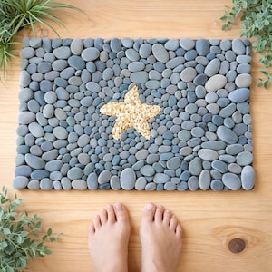 Puede incluir: Una alfombra de baño rectangular hecha de guijarros grises lisos. Una estrella de mar hecha de pequeños guijarros beige está en el centro. La alfombra está sobre un suelo de madera, con dos pies visibles en la parte inferior. Plantas decorativas en el fondo.