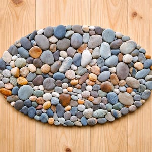 Op de afbeelding: Ovale decoratieve stenen mat bestaande uit rivierstenen van verschillende grootte en kleur. De stenen zijn gerangschikt op een licht houten oppervlak, waardoor een natuurlijk en gestructureerd uiterlijk ontstaat. De stenen zijn in tinten grijs, bruin en blauw.