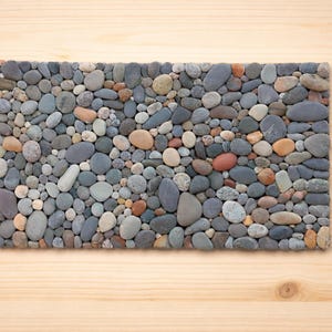 Pu&ograve; includere: Mosaico rettangolare di ciottoli di fiume lisci di varie dimensioni e colori. Le pietre sono grigie, marroni e beige, disposte su una superficie di legno chiaro. Un elemento decorativo.