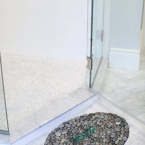May include: Oval-shaped pebble bath mat with a dragonfly design. The mat is made of small, gray stones and features a teal dragonfly mosaic in the center. The mat is placed in a shower with a glass door and white tile.