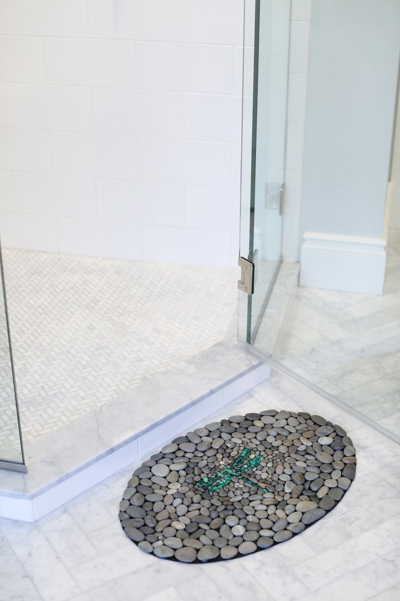 May include: Oval-shaped pebble bath mat with a dragonfly design. The mat is made of small, gray stones and features a teal dragonfly mosaic in the center. The mat is placed in a shower with a glass door and white tile.