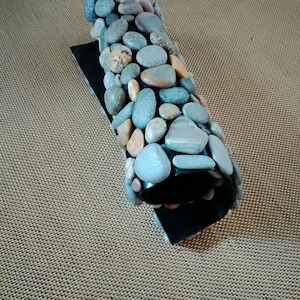 Pu&ograve; includere: Un oggetto decorativo curvo con una base nera e una parte superiore ricoperta di ciottoli di fiume lisci e multicolori. Le pietre sono principalmente sfumature di grigio, blu e marrone.