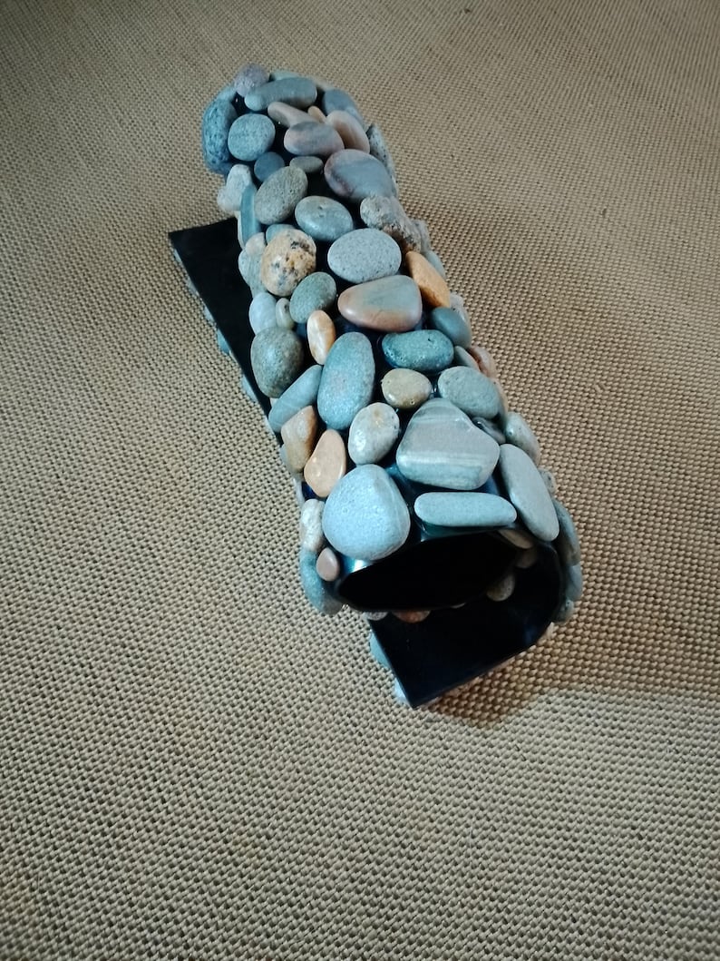 Pu&ograve; includere: Un oggetto decorativo curvo con una base nera e una parte superiore ricoperta di ciottoli di fiume lisci e multicolori. Le pietre sono principalmente sfumature di grigio, blu e marrone.