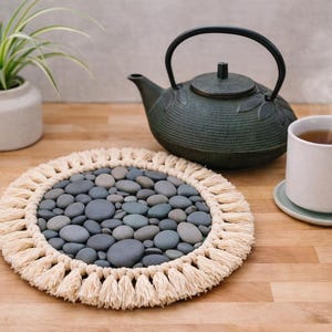 May include: A handmade macrame trivet with a pebble design, a cast iron teapot, and a white mug of tea on a wooden surface. A small potted plant and a saucer are also visible.