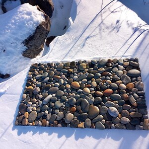 Pu&ograve; includere: Un tappeto rettangolare in pietra composto da ciottoli di fiume lisci di varie dimensioni, nei toni del grigio, marrone e beige. Il tappeto &egrave; posto in un ambiente innevato, con ombre proiettate sulla neve. Le pietre sono ben compattate, creando una superficie strutturata. Circa 60 cm x 40 cm.