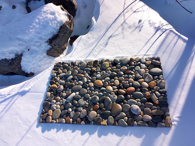Pu&ograve; includere: Un tappeto rettangolare in pietra composto da ciottoli di fiume lisci di varie dimensioni, nei toni del grigio, marrone e beige. Il tappeto &egrave; posto in un ambiente innevato, con ombre proiettate sulla neve. Le pietre sono ben compattate, creando una superficie strutturata. Circa 60 cm x 40 cm.