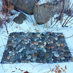 Pu&ograve; includere: Un tappeto di pietra rettangolare per esterni, composto da ciottoli lisci di varie dimensioni, grigi, marroni e beige. Il tappeto &egrave; circondato dalla neve, con rami spogli e grandi rocce sullo sfondo. Design rustico.