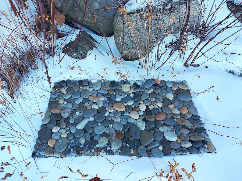 Pu&ograve; includere: Un tappeto di pietra rettangolare per esterni, composto da ciottoli lisci di varie dimensioni, grigi, marroni e beige. Il tappeto &egrave; circondato dalla neve, con rami spogli e grandi rocce sullo sfondo. Design rustico.