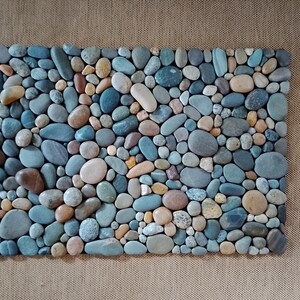 Pu&ograve; includere: Un mosaico rettangolare composto da ciottoli di fiume lisci e colorati. Le pietre variano per dimensioni e tonalit&agrave;, tra cui sfumature di blu, marrone, beige e grigio. La disposizione crea una superficie naturale e strutturata.
