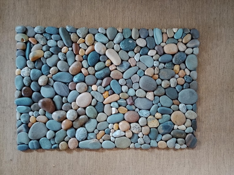 Pu&ograve; includere: Un mosaico rettangolare composto da ciottoli di fiume lisci e colorati. Le pietre variano per dimensioni e tonalit&agrave;, tra cui sfumature di blu, marrone, beige e grigio. La disposizione crea una superficie naturale e strutturata.