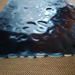 Pu&ograve; includere: Un vassoio rettangolare metallico scuro con una texture martellata. Il vassoio &egrave; sostenuto da piccole pietre arrotondate lungo il bordo inferiore. La superficie riflette la luce.
