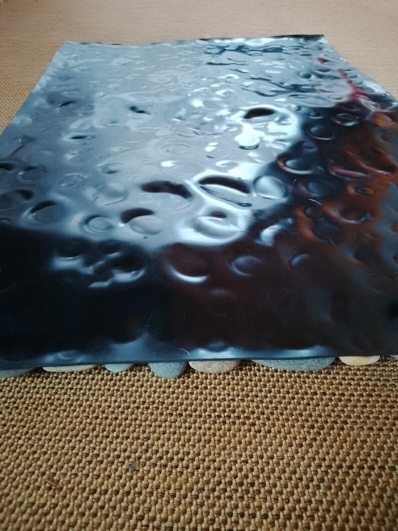 Pu&ograve; includere: Un vassoio rettangolare metallico scuro con una texture martellata. Il vassoio &egrave; sostenuto da piccole pietre arrotondate lungo il bordo inferiore. La superficie riflette la luce.