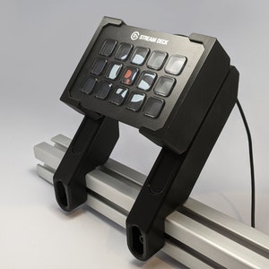 May include: A black Stream Deck with 15 buttons, mounted on a silver metal rail system. The Stream Deck is connected to a computer via a black cable.