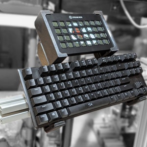 May include: A black keyboard with a Stream Deck mounted on top. The keyboard has a silver metal bar attached to the side. The Stream Deck has a grid of buttons with a red and white circle in the center.