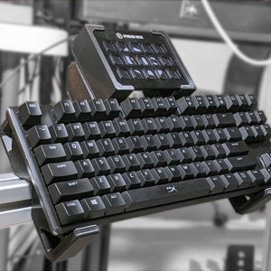 Puede incluir: Un teclado negro con un Stream Deck montado en la parte superior. El teclado tiene un diseño en blanco y negro con un logotipo de HyperX en la esquina inferior derecha. El Stream Deck tiene un diseño en negro y gris con un logotipo de Stream Deck en la parte superior.
