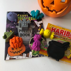 May include: A collection of Halloween-themed decorations and treats. Includes an orange jack-o'-lantern, a puzzle book, and various plastic figures like a bat, pumpkin, witch hat, and cauldron. A package of Haribo candies is also present.