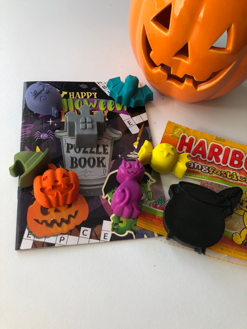 May include: A collection of Halloween-themed decorations and treats. Includes an orange jack-o'-lantern, a puzzle book, and various plastic figures like a bat, pumpkin, witch hat, and cauldron. A package of Haribo candies is also present.