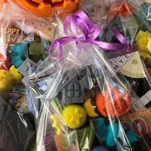May include: A collection of Halloween-themed wax melts in various shapes and colors, including pumpkins, bats, and stars, packaged in clear cellophane bags and tied with a purple ribbon. An orange pumpkin-shaped container is in the background.