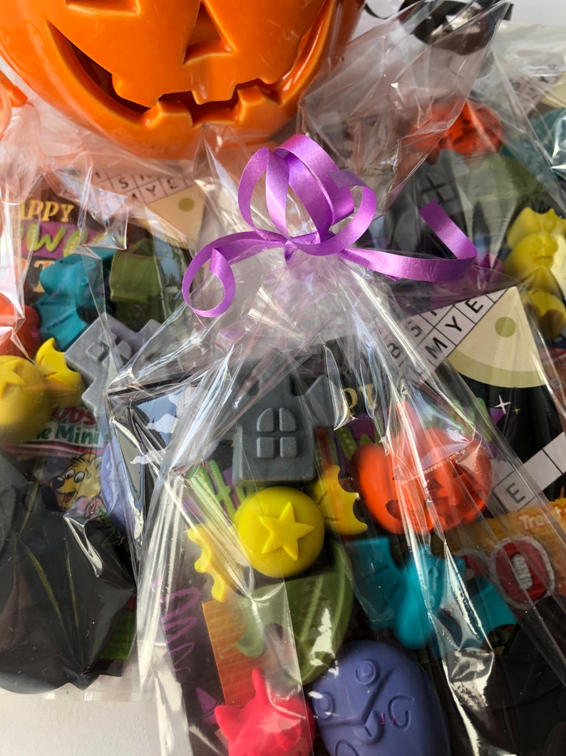 May include: A collection of Halloween-themed wax melts in various shapes and colors, including pumpkins, bats, and stars, packaged in clear cellophane bags and tied with a purple ribbon. An orange pumpkin-shaped container is in the background.