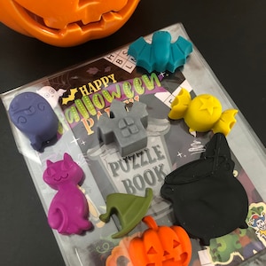 May include: A collection of Halloween-themed rubber charms in various shapes and colors, including a bat, cat, pumpkin, and cauldron. The charms are arranged on a clear plastic package with a "Happy Halloween Party" puzzle book.