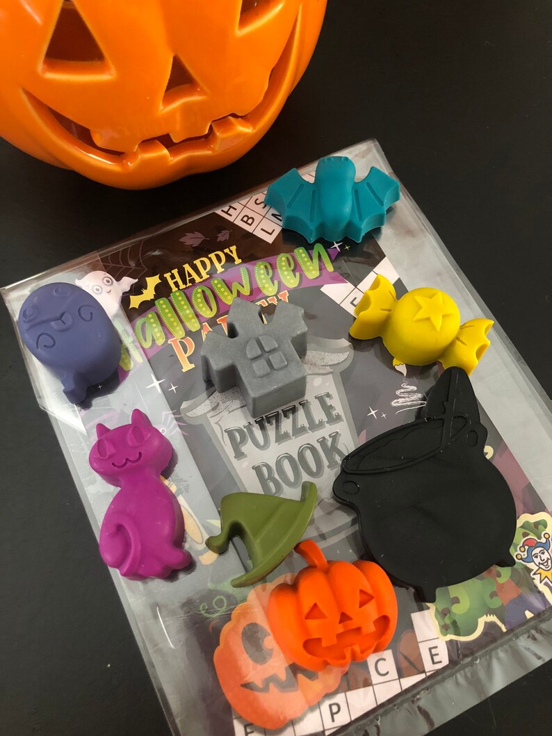 May include: A collection of Halloween-themed rubber charms in various shapes and colors, including a bat, cat, pumpkin, and cauldron. The charms are arranged on a clear plastic package with a "Happy Halloween Party" puzzle book.