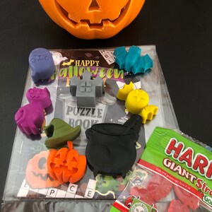 May include: An orange ceramic jack-o'-lantern sits above a Halloween puzzle book and various Halloween-themed wax melts. The melts include a ghost, cat, witch hat, pumpkin, cauldron, and bat. A bag of strawberry-flavored gummy candies is also present.