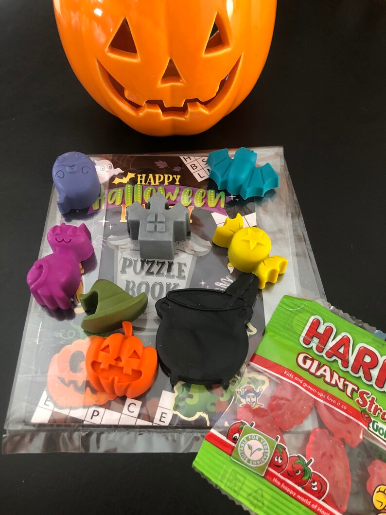 May include: An orange ceramic jack-o'-lantern sits above a Halloween puzzle book and various Halloween-themed wax melts. The melts include a ghost, cat, witch hat, pumpkin, cauldron, and bat. A bag of strawberry-flavored gummy candies is also present.