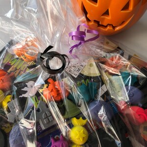 May include: A Halloween-themed gift basket wrapped in clear cellophane. The basket contains various Halloween-shaped candies and decorations, including pumpkins, ghosts, and cats. An orange ceramic jack-o'-lantern sits on top, with purple and black ribbons for decoration.