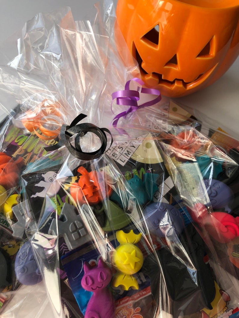 May include: A Halloween-themed gift basket wrapped in clear cellophane. The basket contains various Halloween-shaped candies and decorations, including pumpkins, ghosts, and cats. An orange ceramic jack-o'-lantern sits on top, with purple and black ribbons for decoration.
