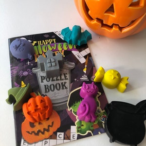 May include: A collection of Halloween-themed decorations, including a jack-o'-lantern, a puzzle book, and various small figurines like a bat, cat, and witch's hat. The decorations are in shades of orange, purple, green, and yellow.
