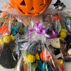 May include: An orange ceramic jack-o'-lantern candy bowl filled with wrapped Halloween-themed treats. The bowl has a carved face and is wrapped in clear cellophane with a purple ribbon. Assorted candies and small toys are visible.