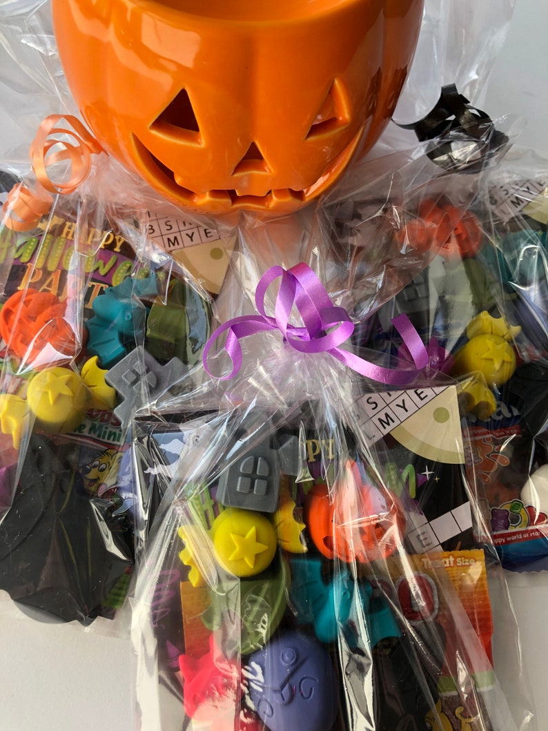 May include: An orange ceramic jack-o'-lantern candy bowl filled with wrapped Halloween-themed treats. The bowl has a carved face and is wrapped in clear cellophane with a purple ribbon. Assorted candies and small toys are visible.