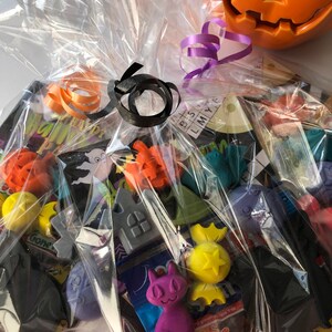 May include: A cellophane-wrapped Halloween gift basket filled with assorted candies and decorative items. The basket is tied with orange, black, and purple ribbons. A carved orange pumpkin sits in the background.