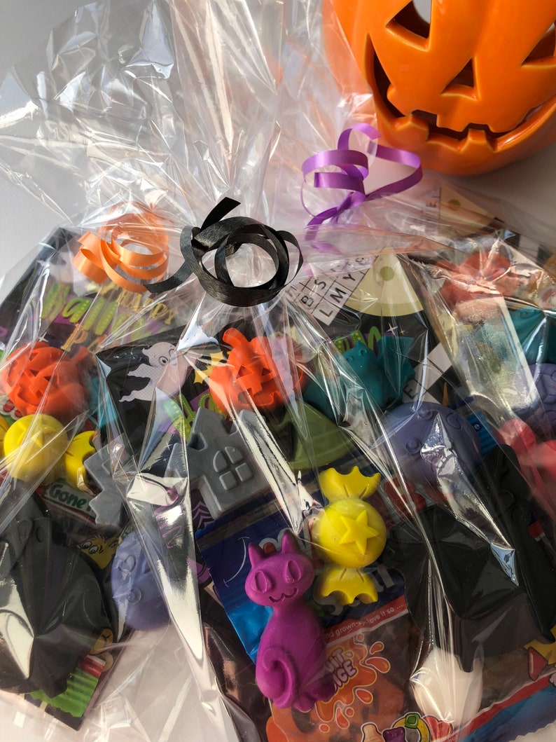 May include: A cellophane-wrapped Halloween gift basket filled with assorted candies and decorative items. The basket is tied with orange, black, and purple ribbons. A carved orange pumpkin sits in the background.